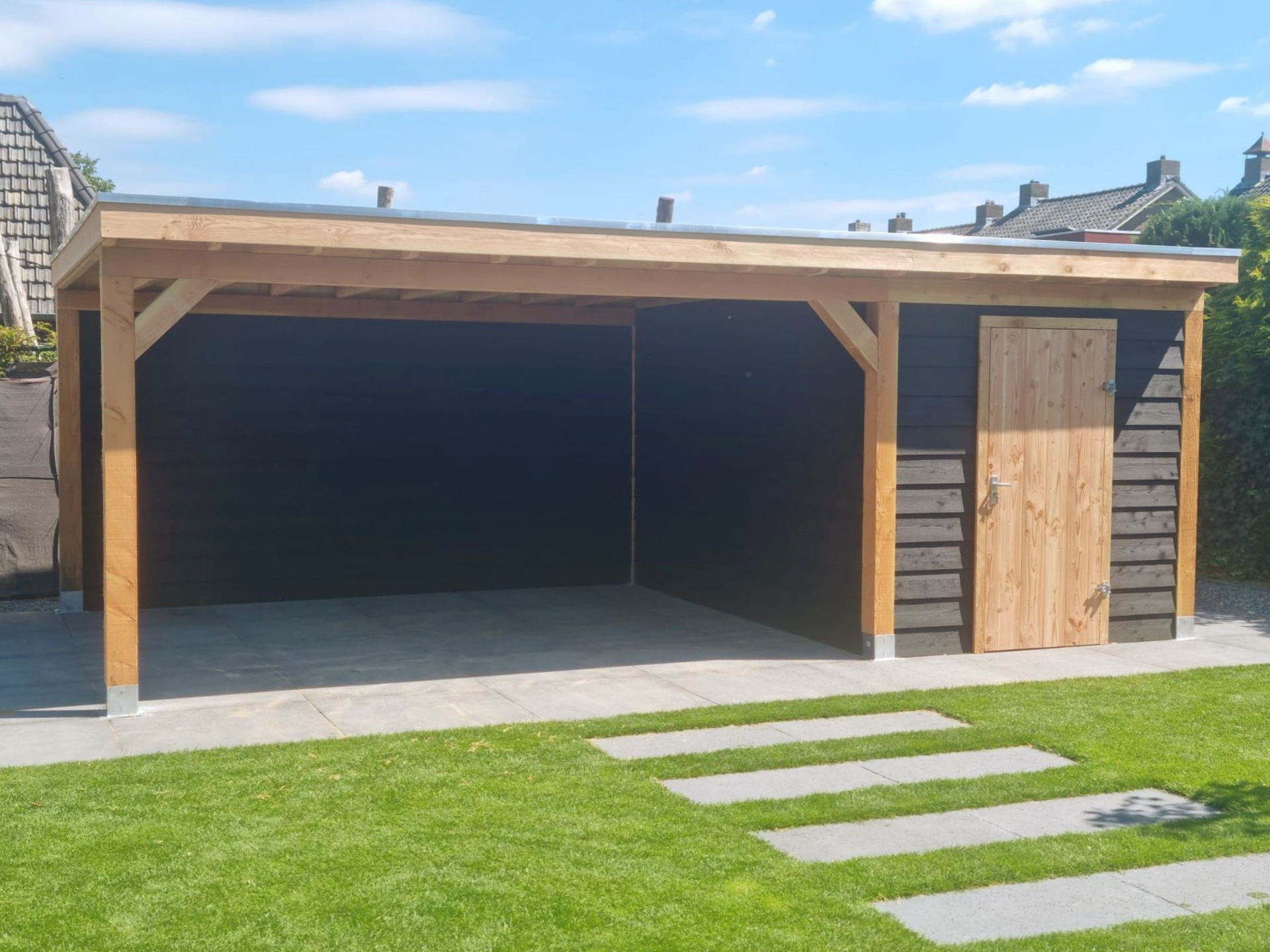 Houten carport met berging, zwarte muren, beige palen en deur, gelegen op een betonnen terras met gazon.