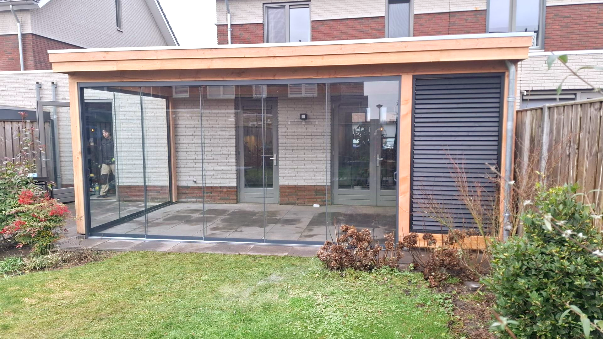 Een houten terrasoverkapping met glazen wanden en een lamellen privacypaneel, in een achtertuin met groen gras.