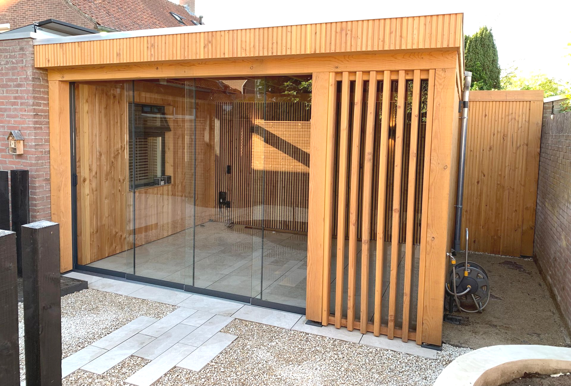 Houten schuur met glazen schuifdeuren en een houten lamellenpaneel aan de rechterkant. Terras met grind.