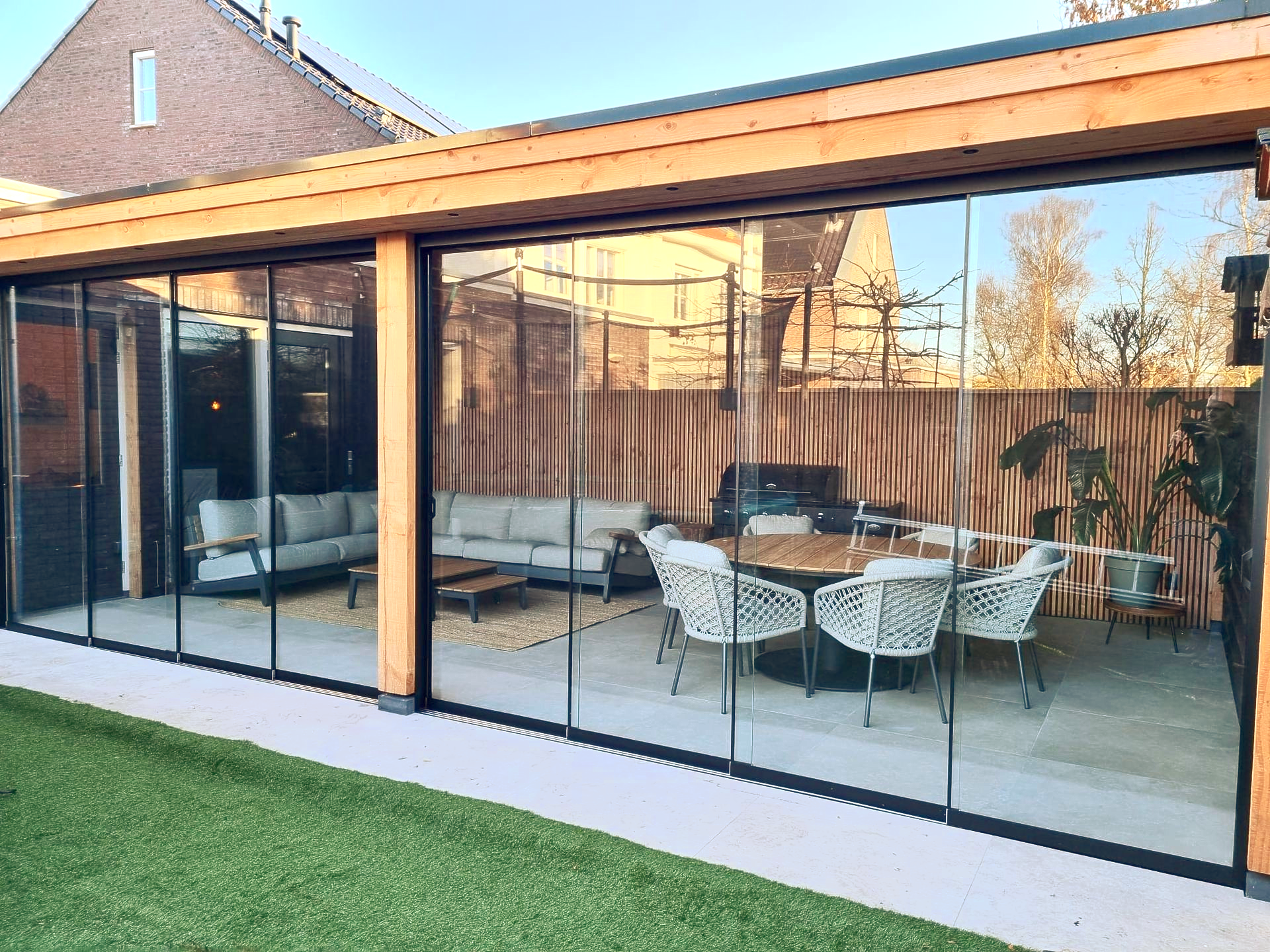 Een patio met glazen wanden en zitgelegenheid buiten, die grenst aan een gebouw met een gazon.