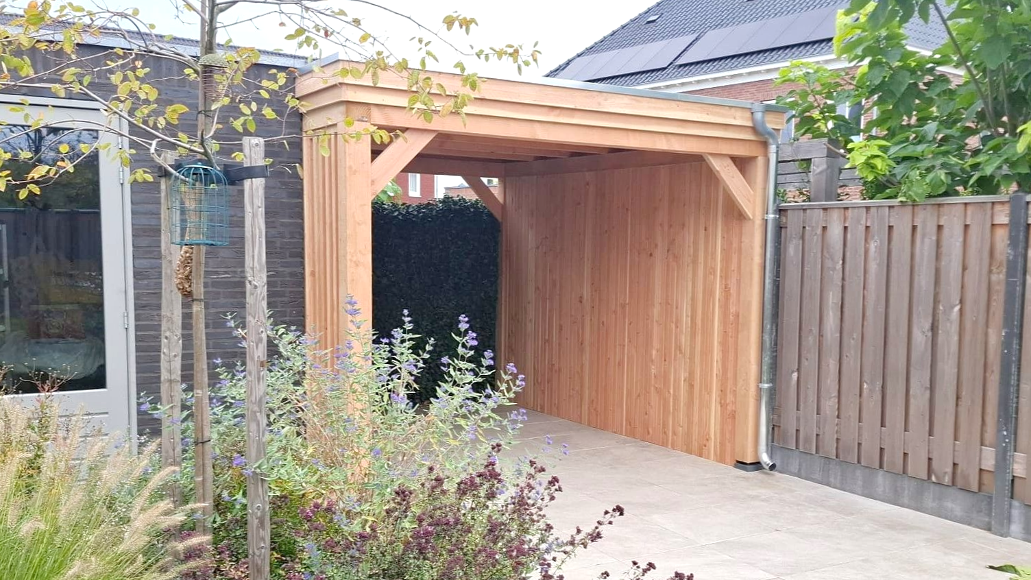 Een houten carport, aangebouwd aan een bakstenen gebouw. ​​De carport beschermt een betonnen oprit.