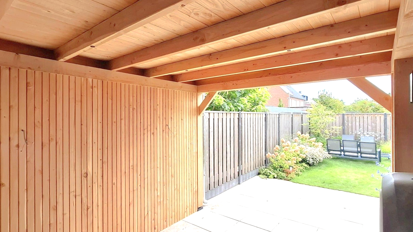 Houten terrasoverkapping met verticale houten panelen, met uitzicht op een omheinde achtertuin met gazon en tuinstoelen.