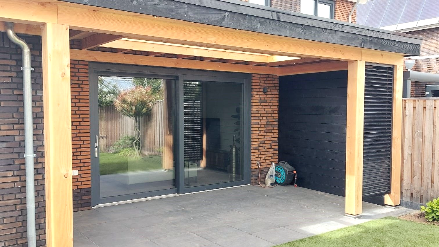 Overdekt terras met schuifdeuren van glas, een bakstenen muur, houten balken en zwarte lambrisering.