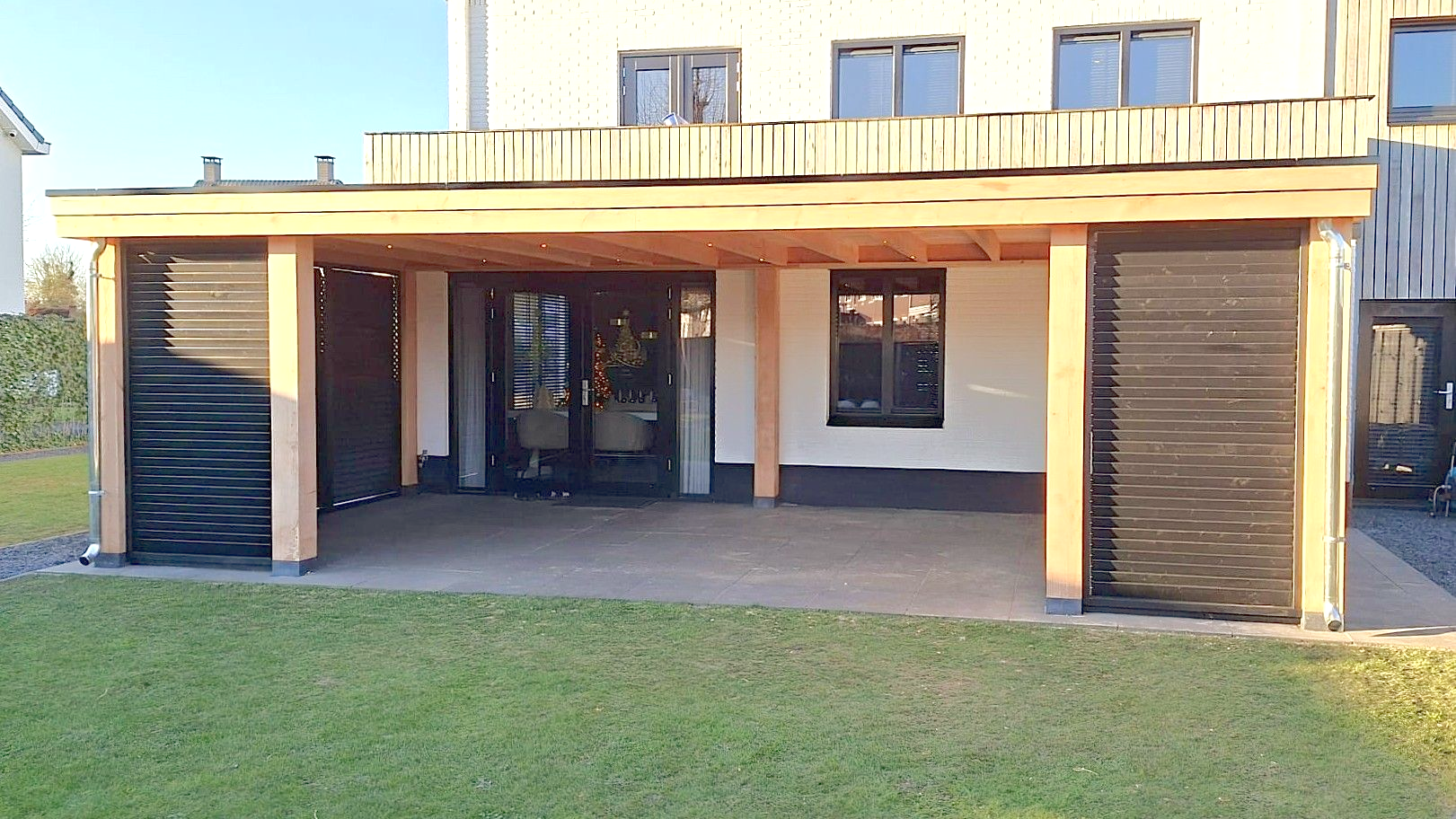 Carport met houten frame, zwarte lattenwanden en betonnen vloer, grenzend aan een huis.