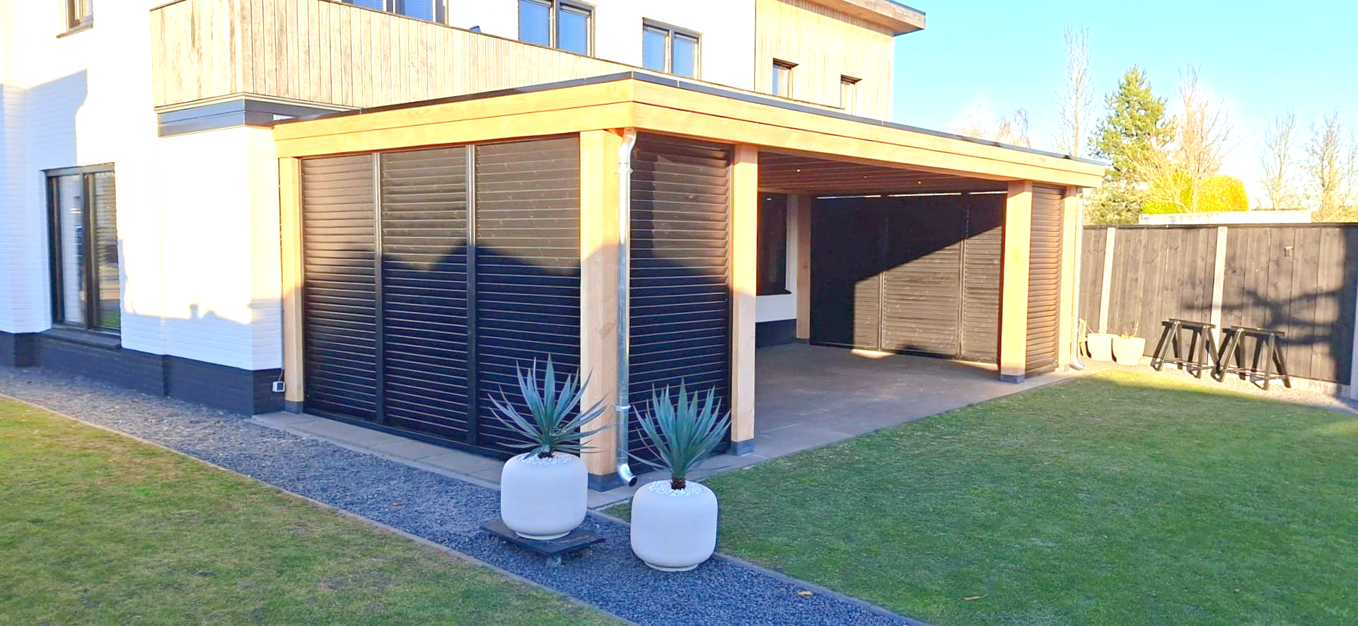 Exterieurbeeld van een moderne carport met zwarte accenten, lichte houten pilaren en een aangelegde tuin met potplanten.