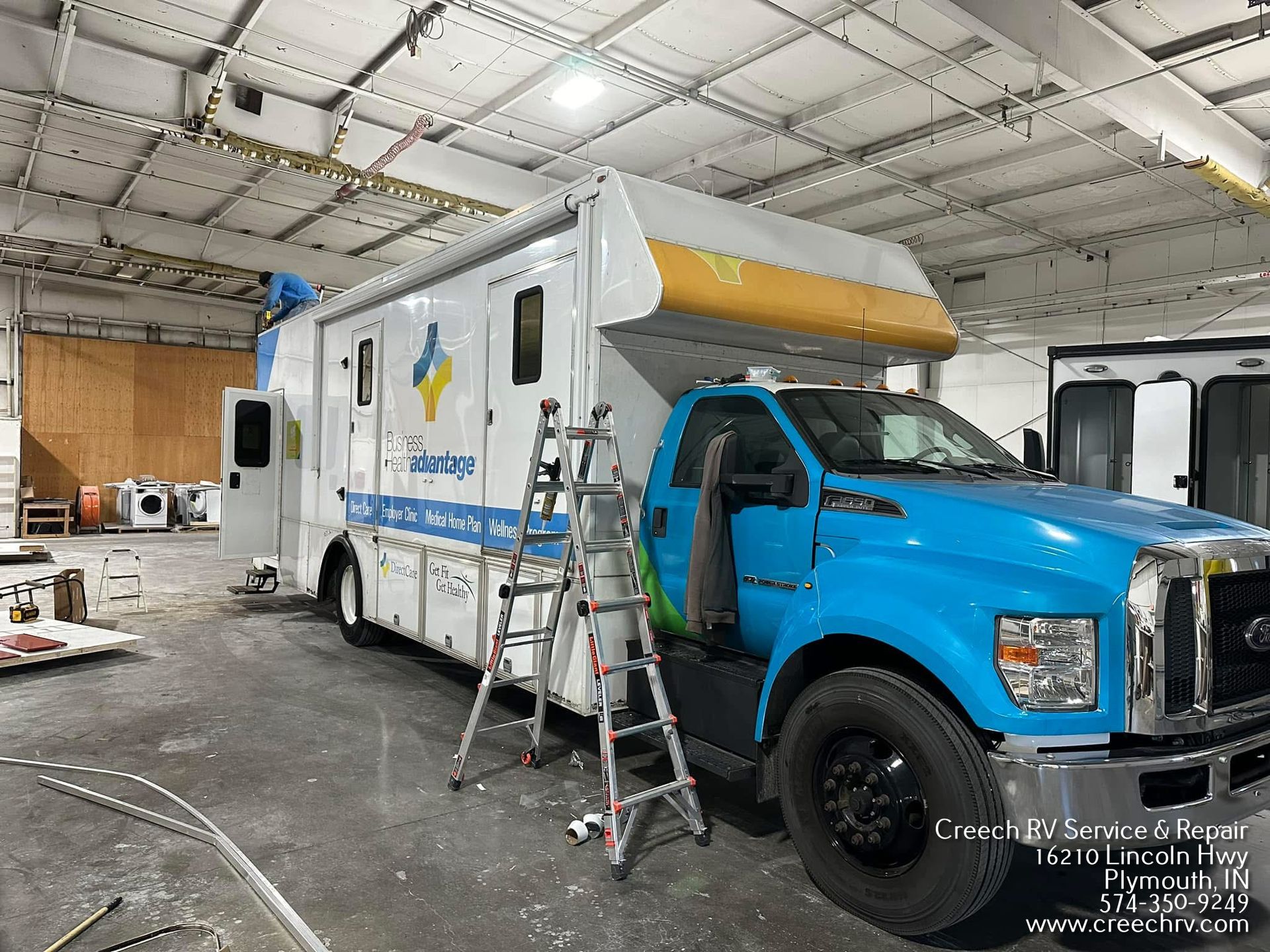 Blue utility truck with ladder in a warehouse garage, next to a large white service vehicle