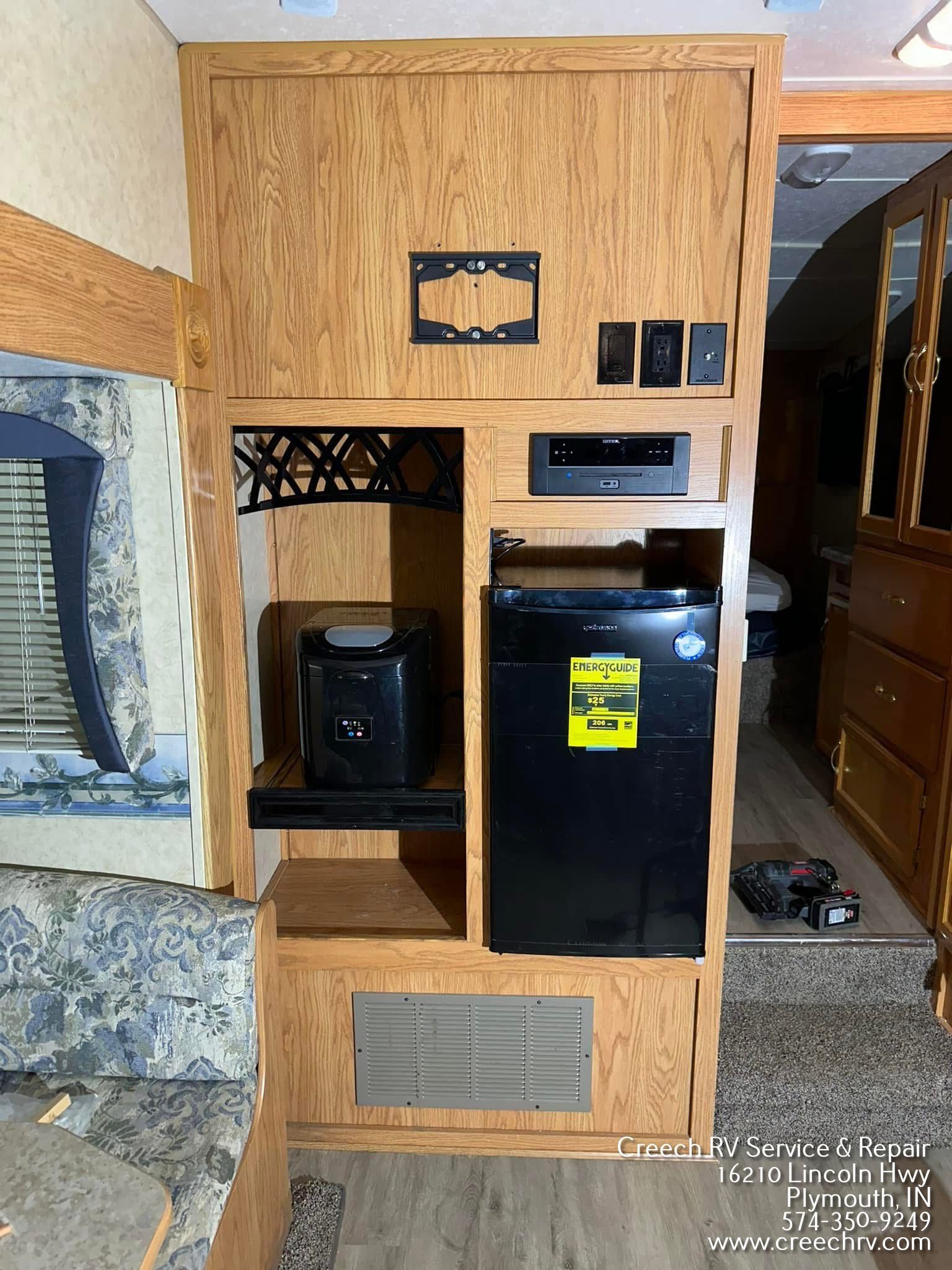 RV interior with wood paneling, featuring a small refrigerator, shelf, and TV mounting bracket.