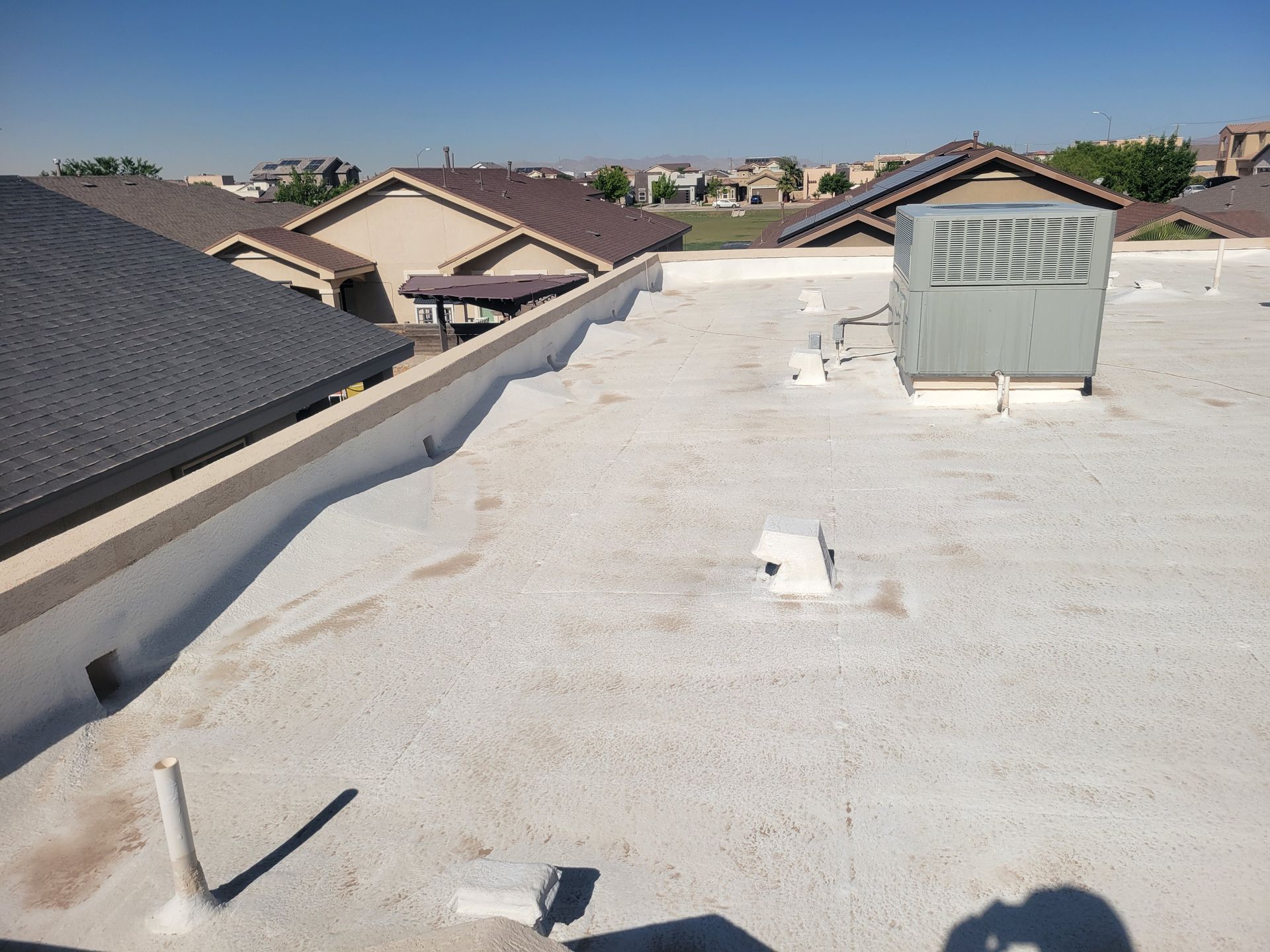 Flat white rooftop with HVAC unit, vents, and neighboring houses under a clear sky.