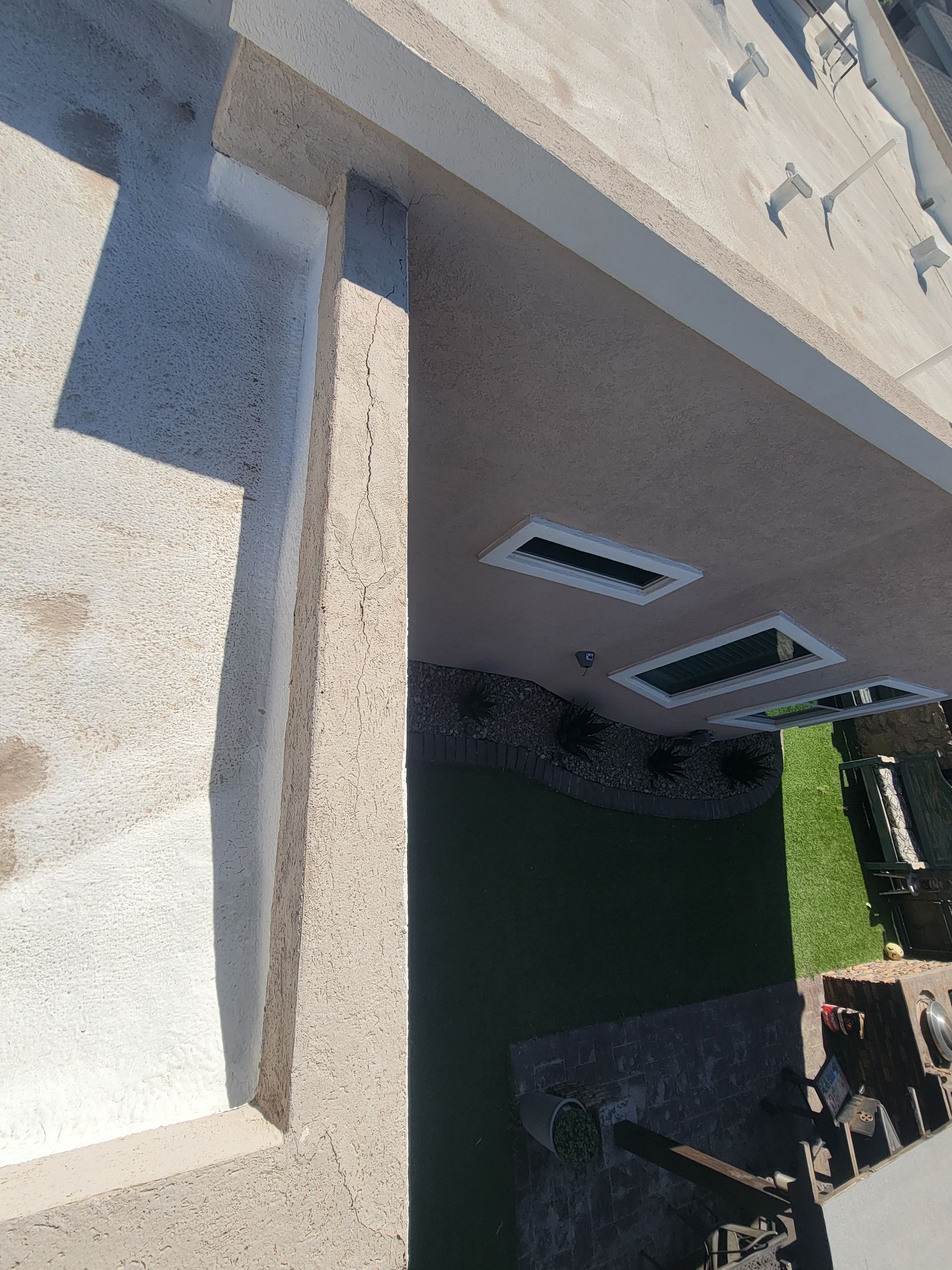 Top-down view of a building roof and wall with two vents above a grassy yard and patio area.