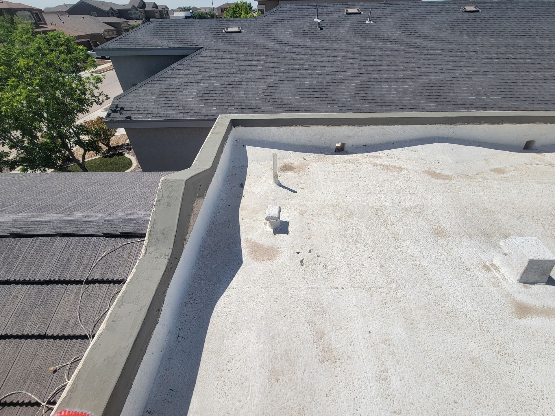 Flat white rooftop beside dark shingled roofs and trees under a bright sky
