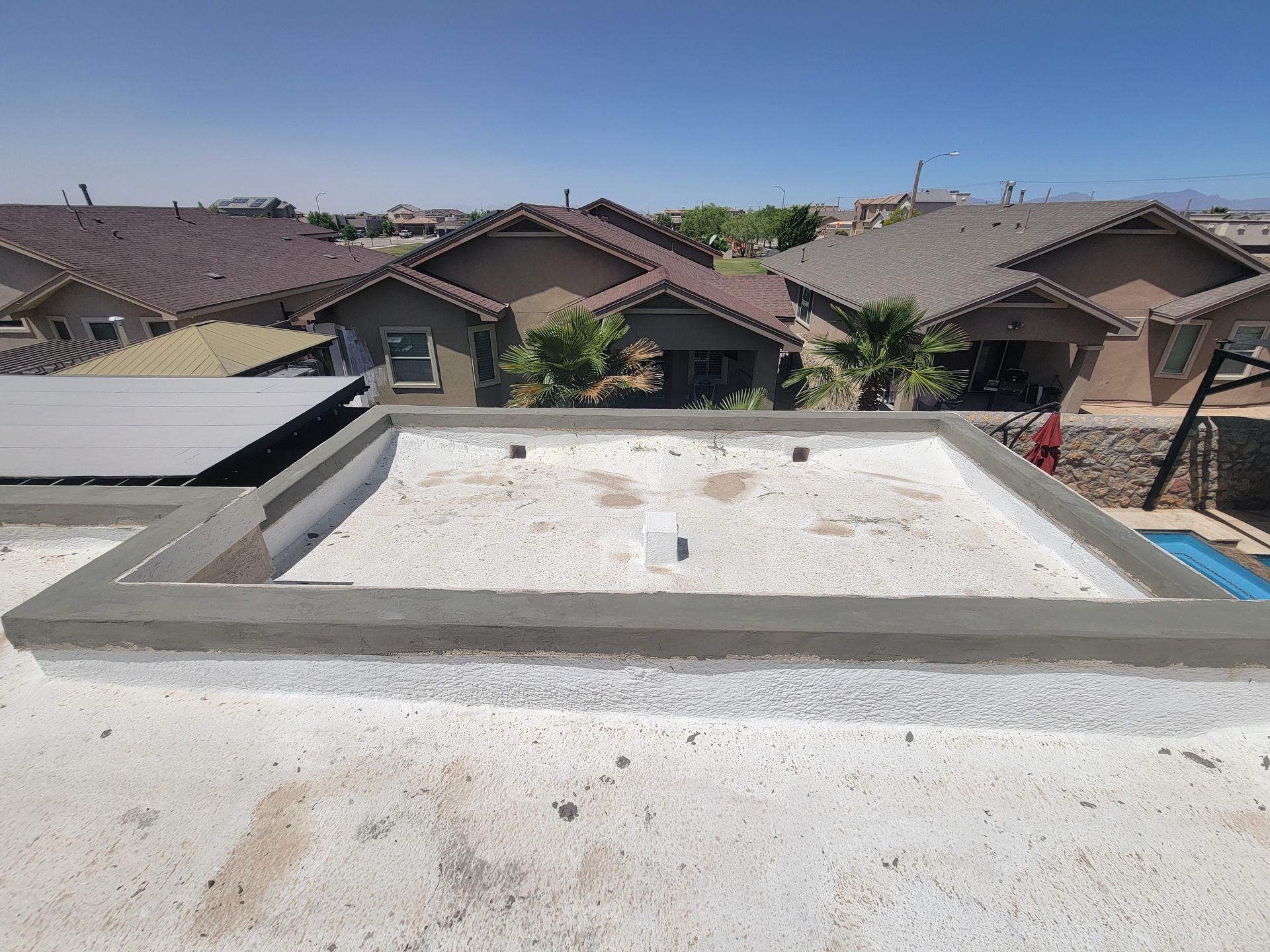 Flat rooftop with white surface and HVAC units, overlooking suburban houses under a clear blue sky