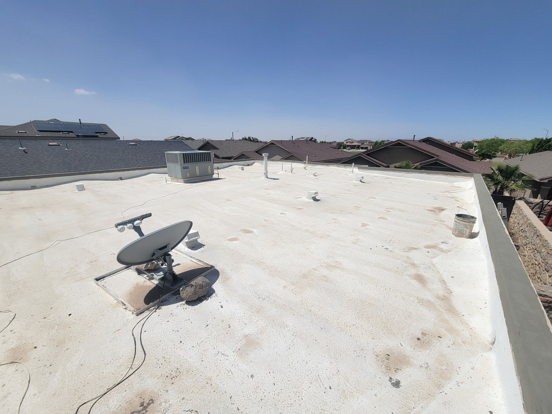 Rooftop with white coating, satellite dish, vents, and neighboring houses under a clear blue sky