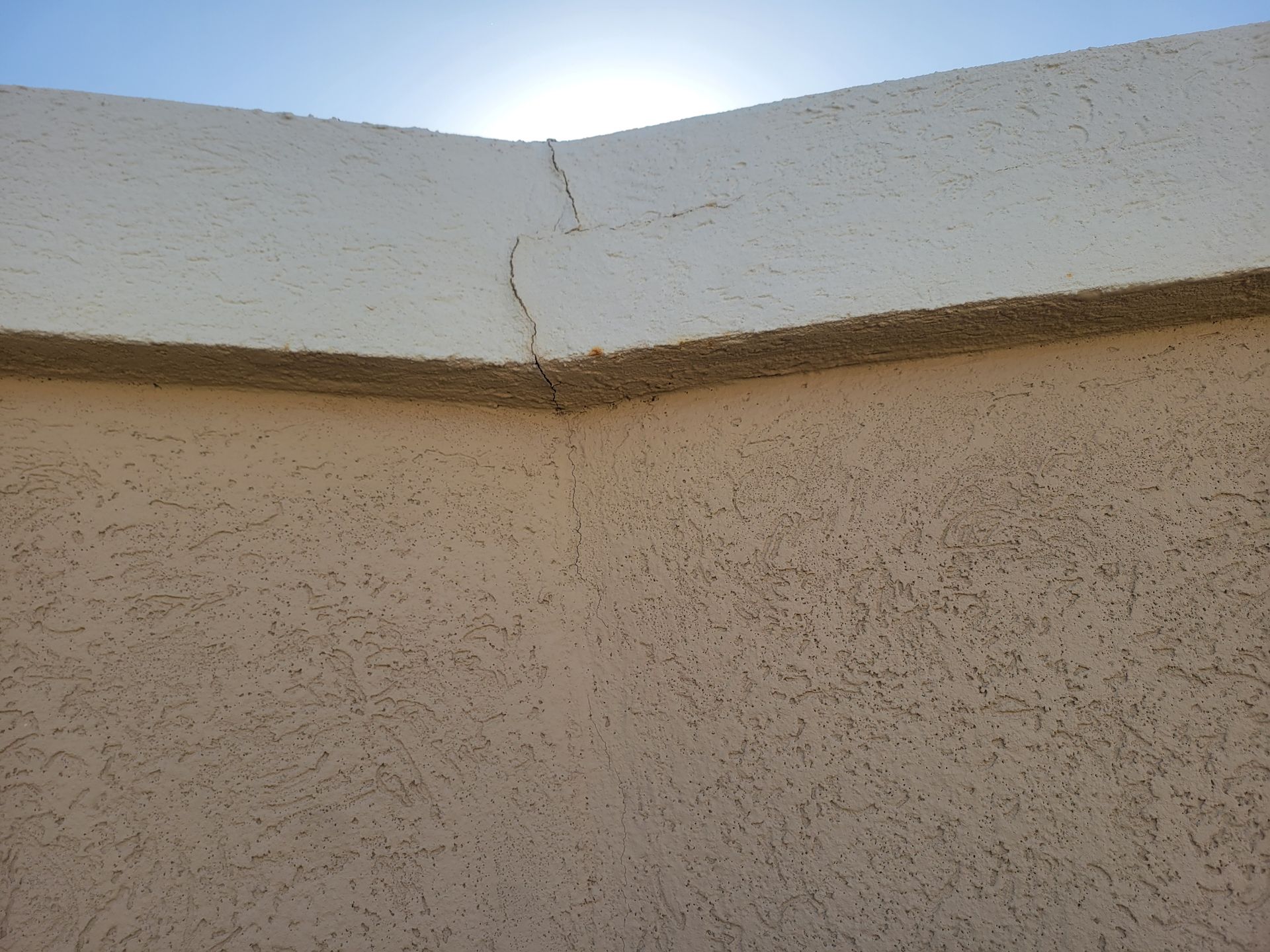 White stucco wall with a diagonal crack at the top, lit by sunlight against a blue sky.