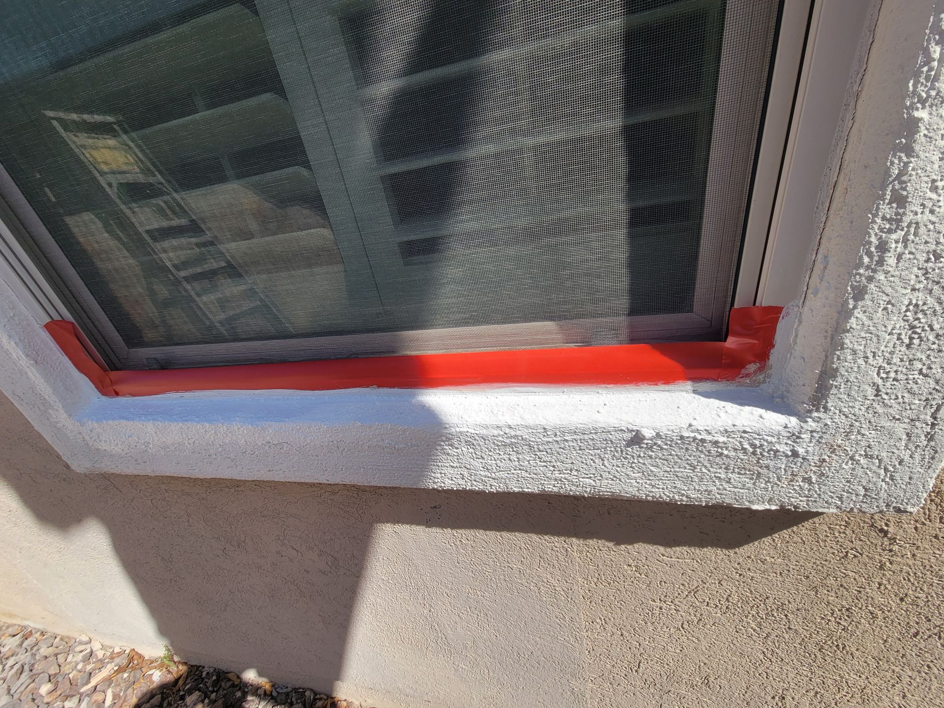 Close-up of a window frame with red sealant around the bottom edge and textured stucco wall.