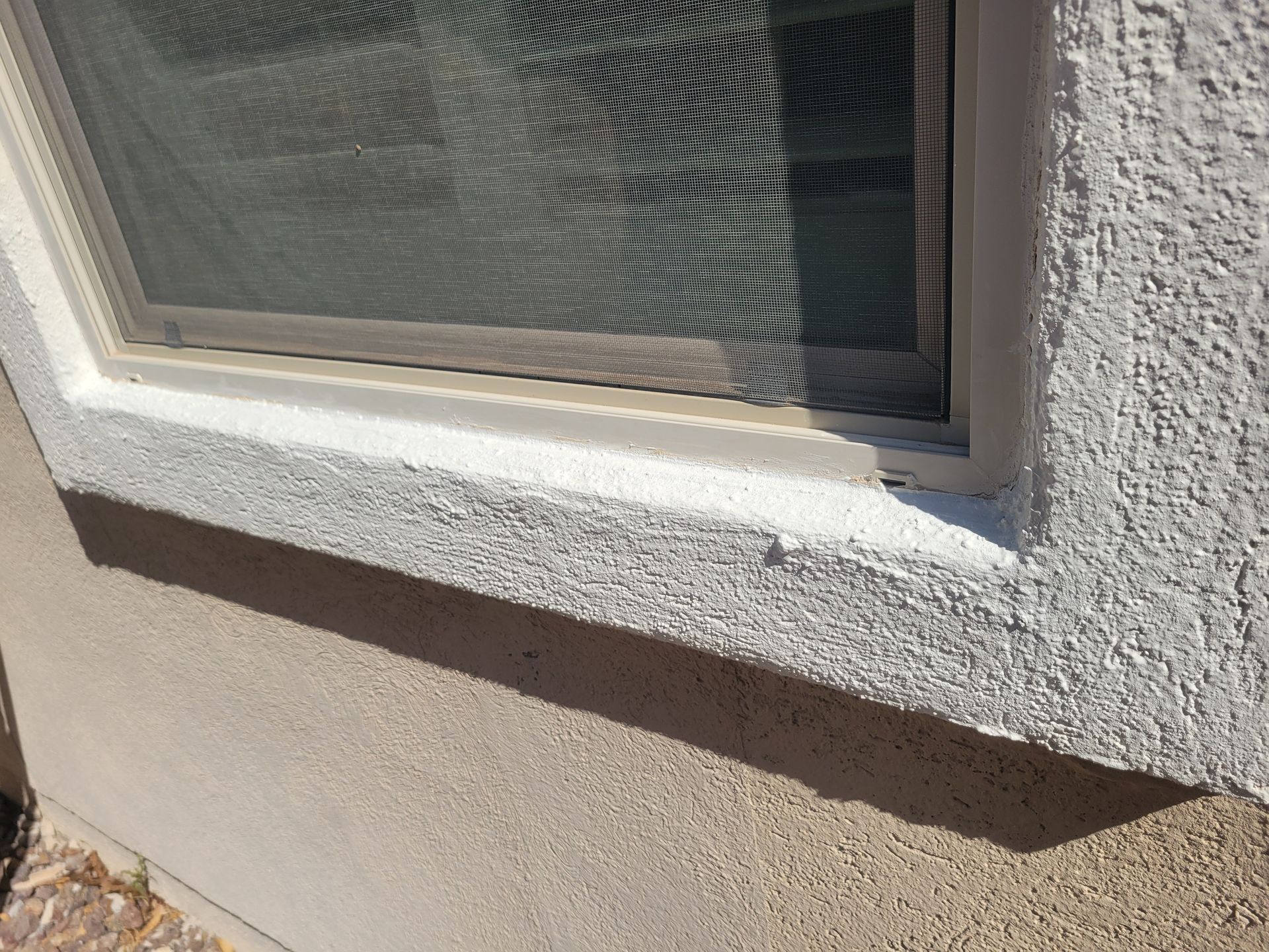 Close-up of a window with a textured white sill and beige stucco wall in sunlight