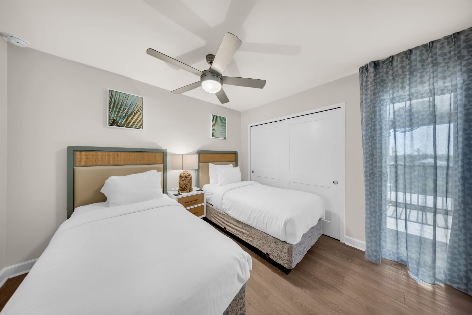 Bright bedroom with two twin beds, ceiling fan, and sliding glass door with blue curtains.