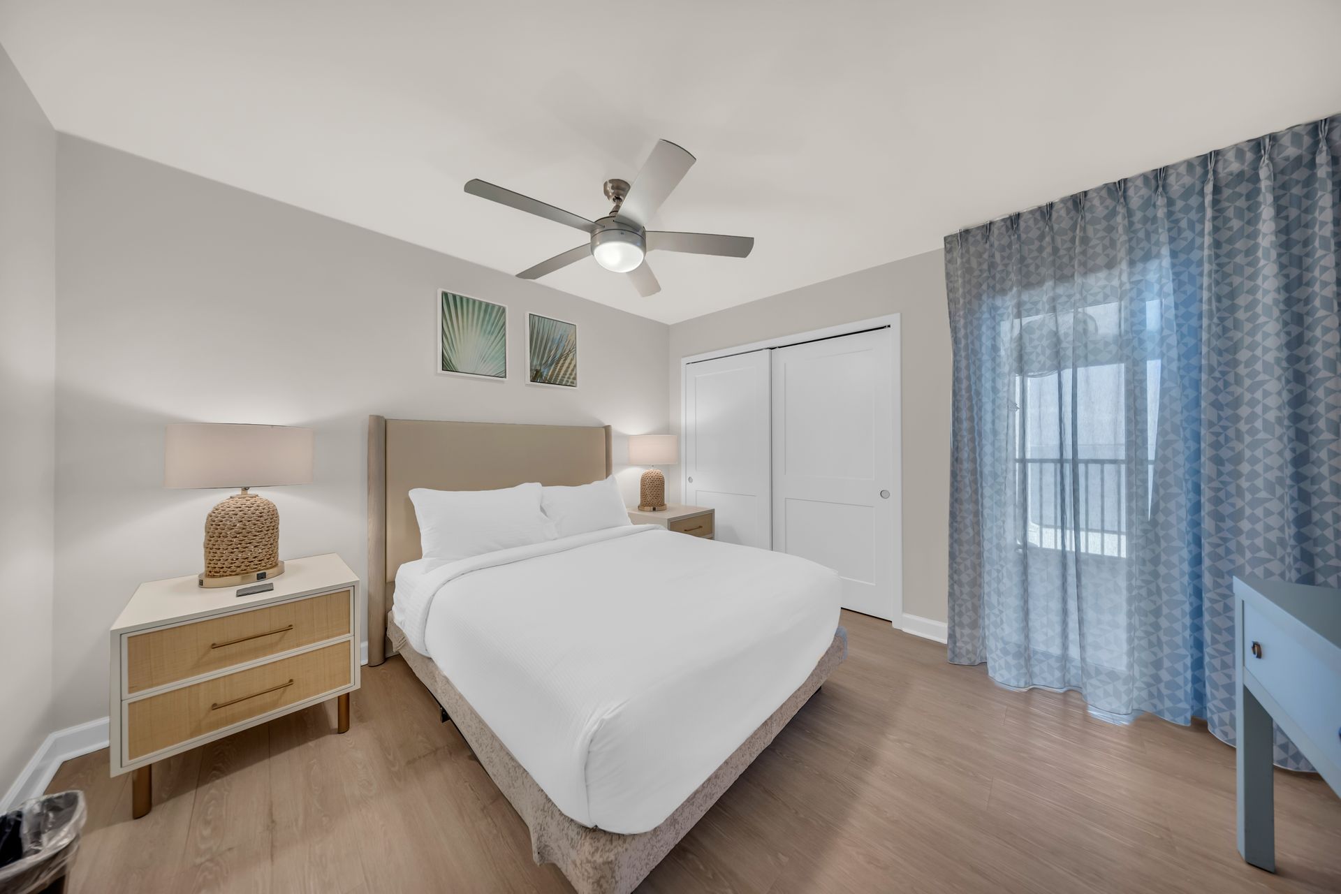 Bright bedroom with white bed, wood furniture, ceiling fan, closet, and blue curtains