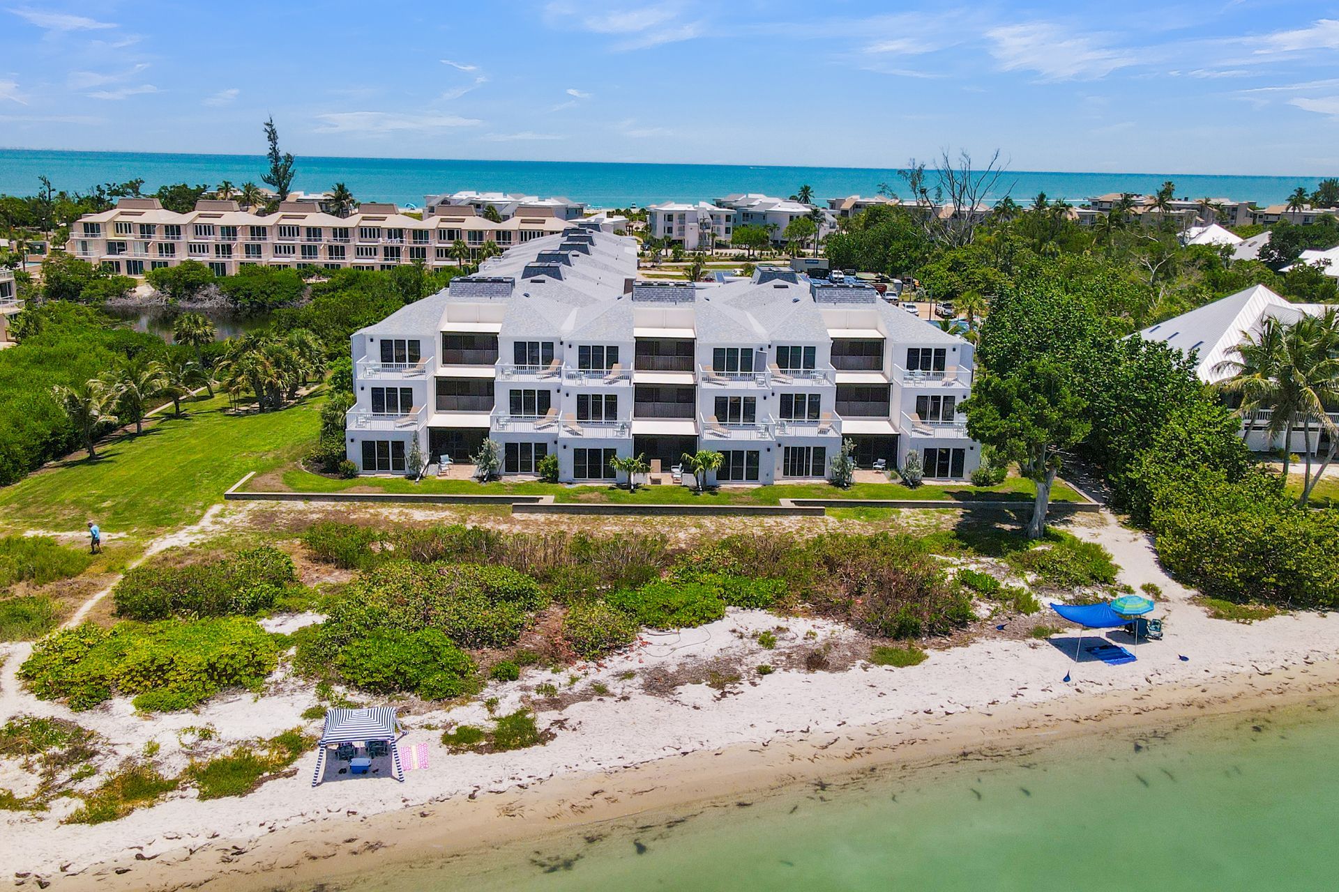 White beachfront condo complex with turquoise water, green landscaping, and sandy shore under a blue sky