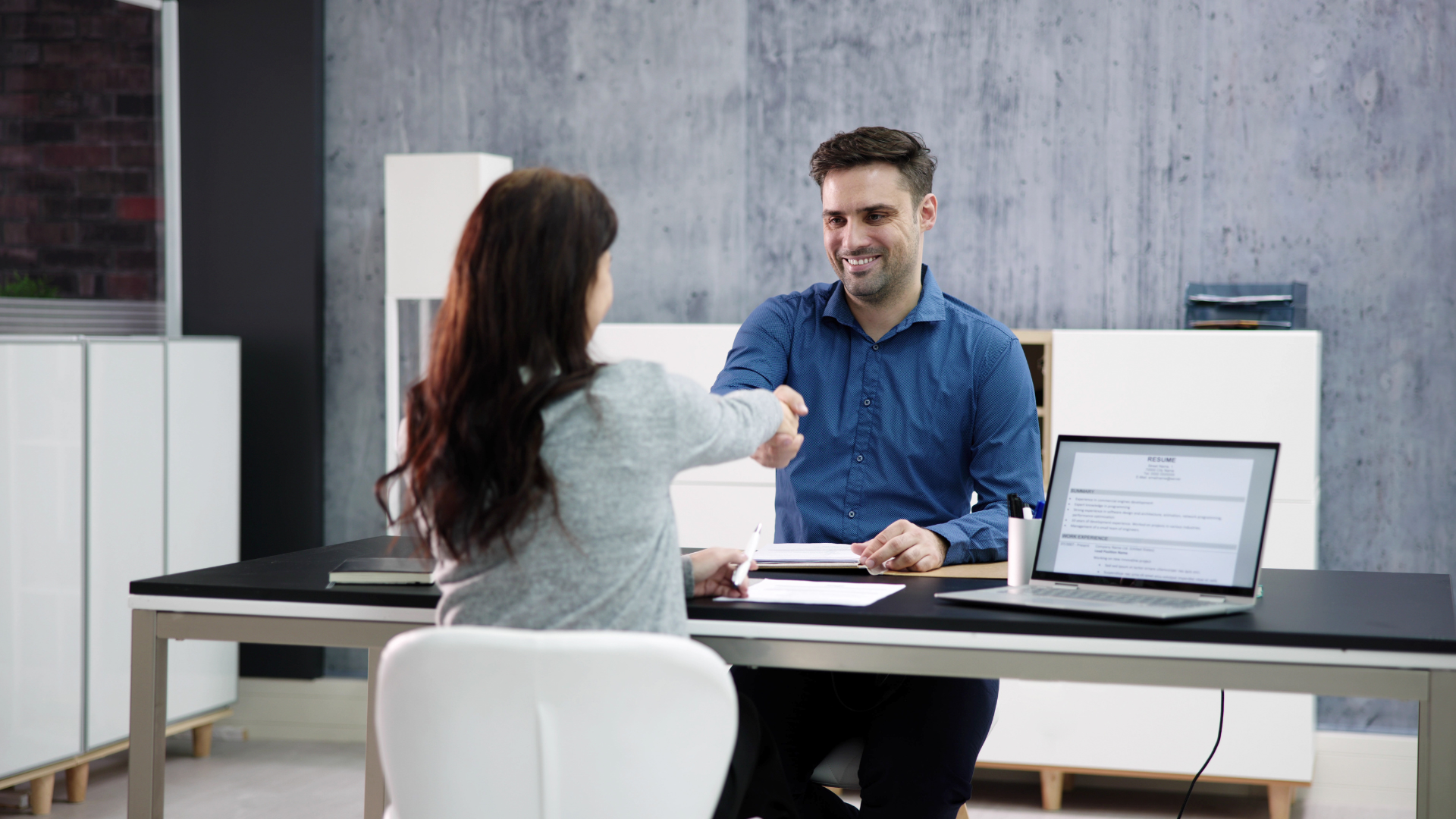 Woman and man shaking hands at a desk; office setting, smiling, blue shirt, laptop.