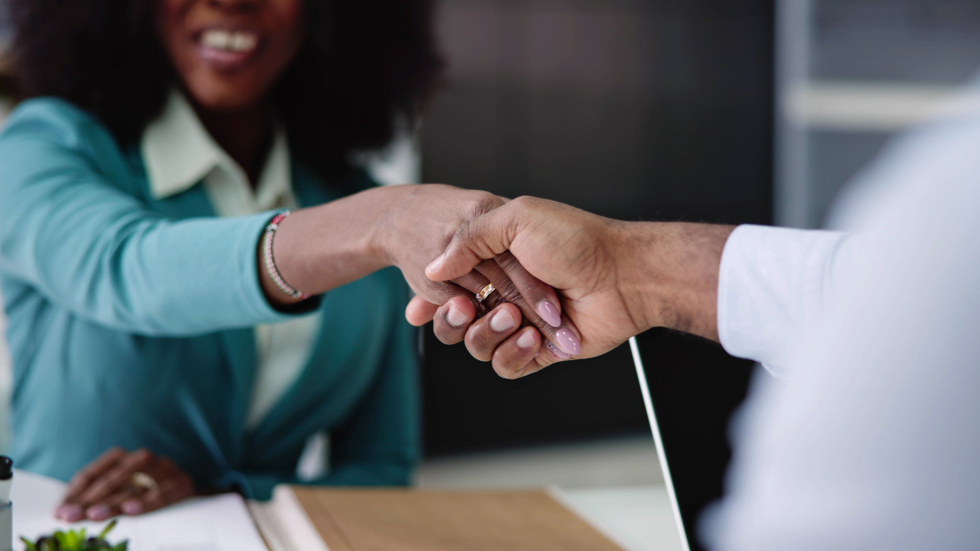 Woman in teal blazer shaking hands with a person in a white shirt at a desk.