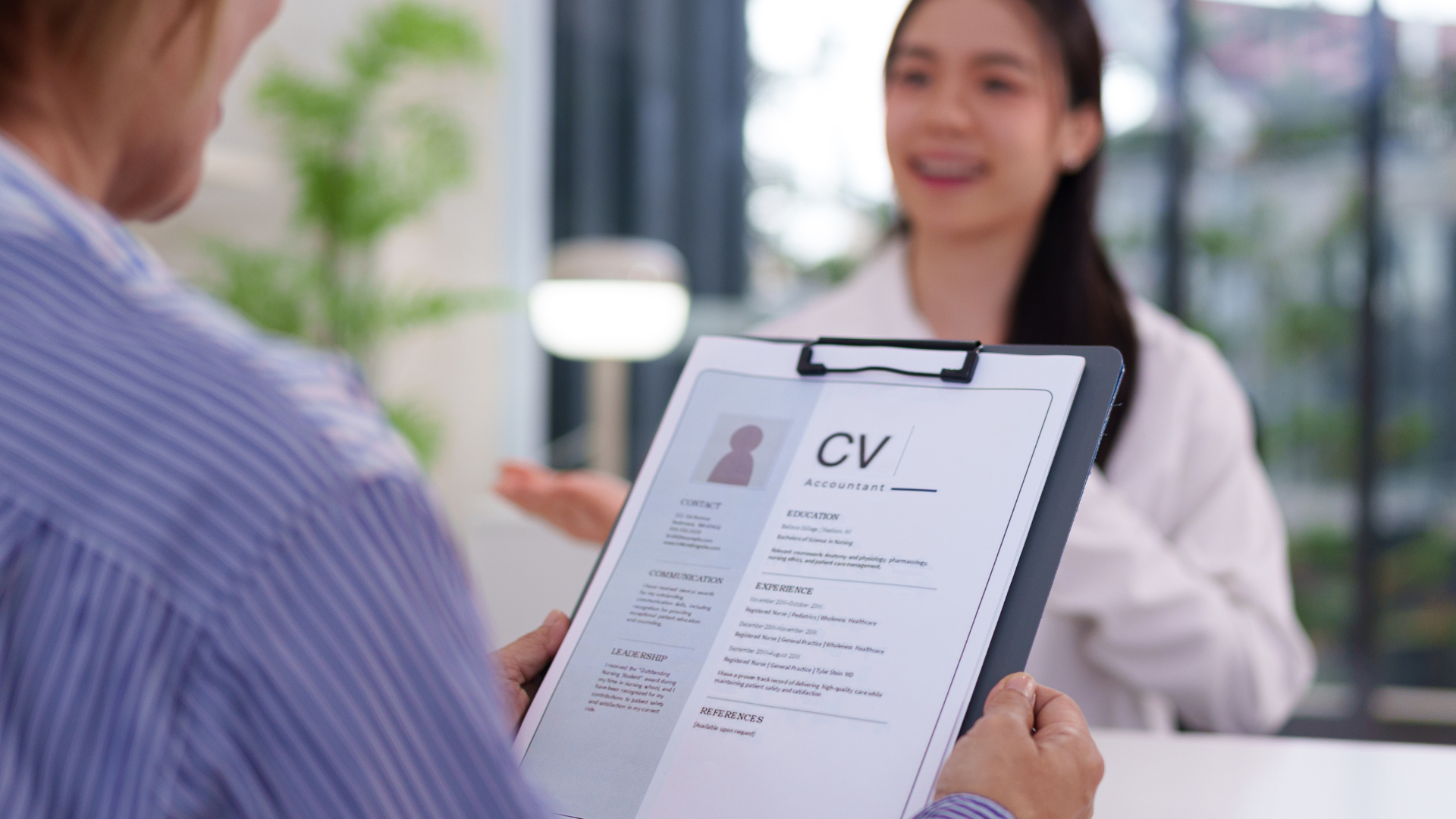 Person holding a CV clipboard, interviewing a smiling woman. Office setting, natural light.