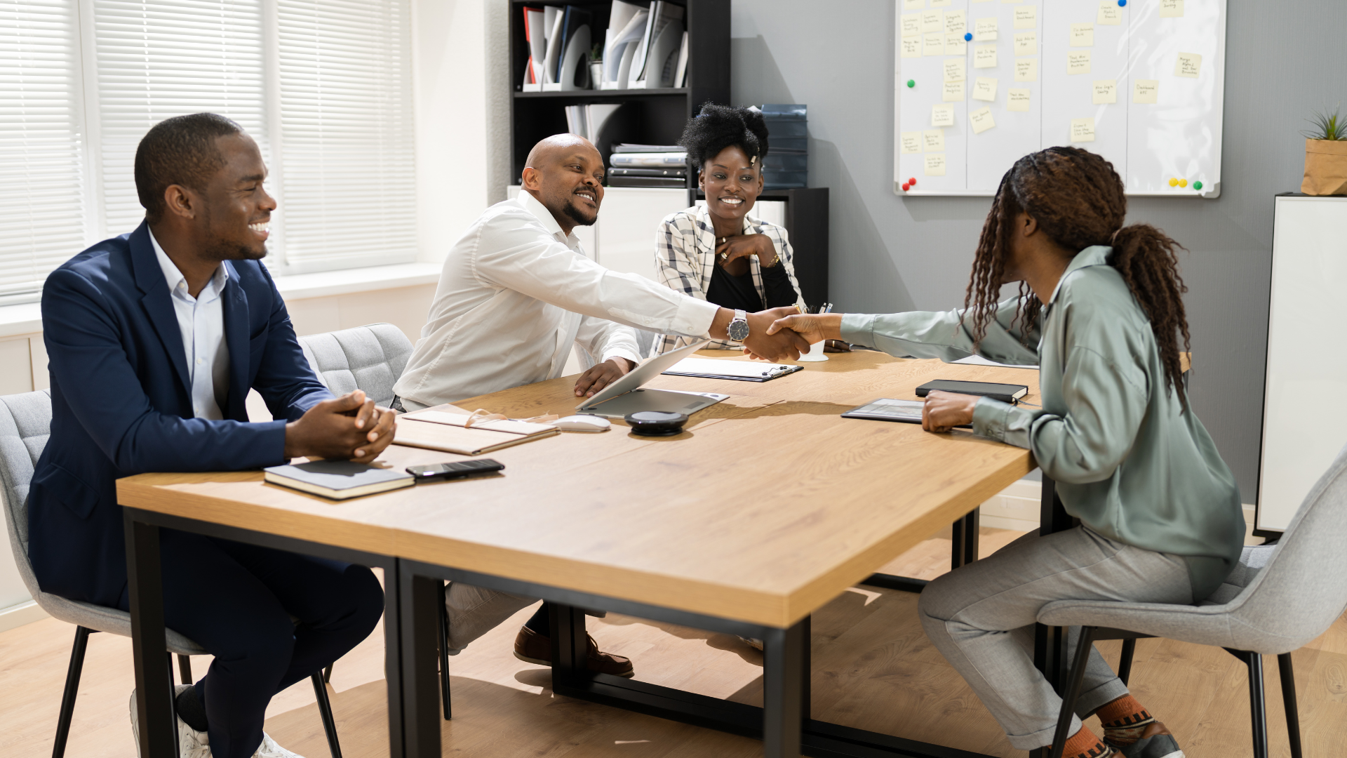 Four people in a conference room, two shaking hands. Smiling faces around a wooden table, near a whiteboard.