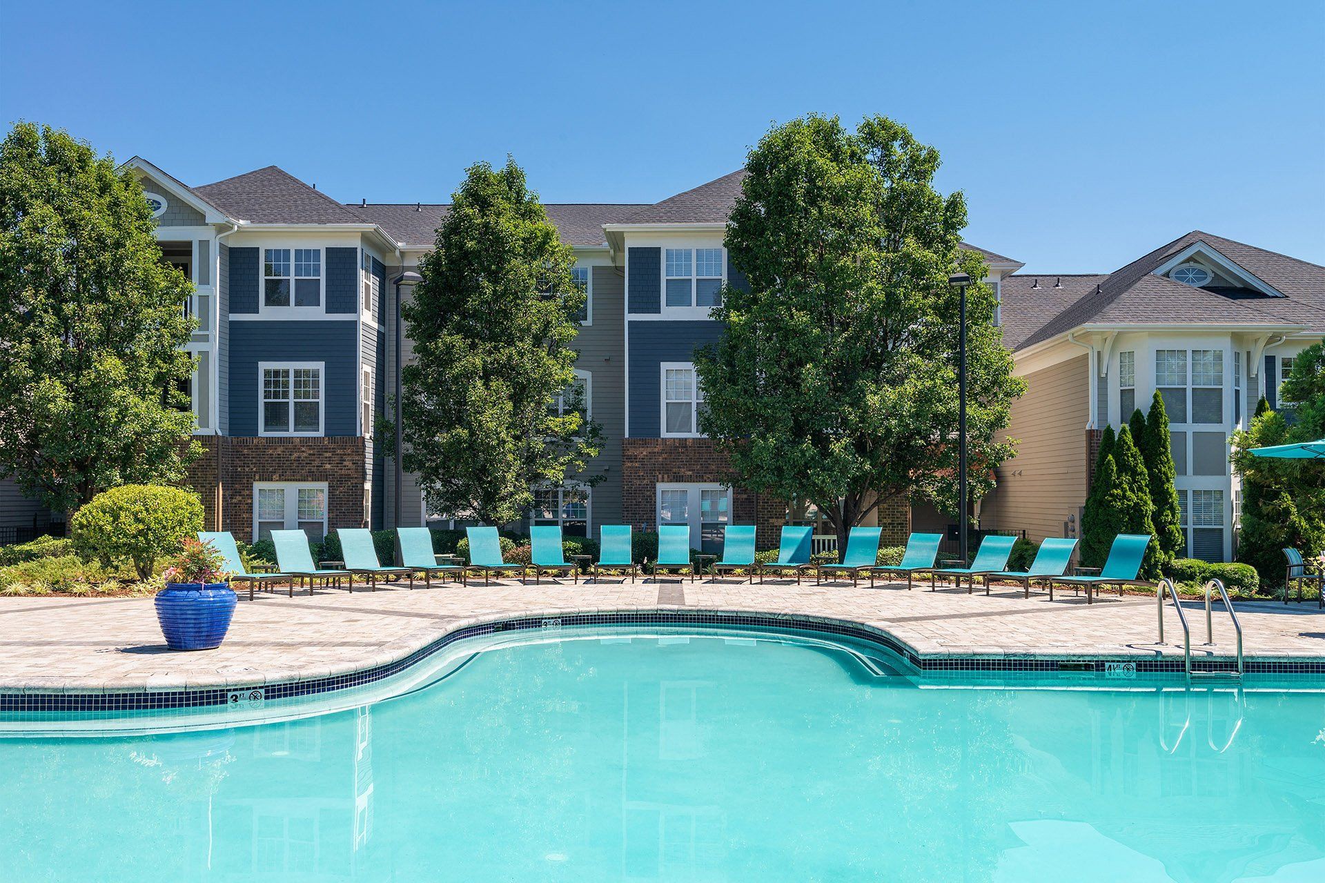 Chapel Hill, NC, Luxury Apartments with In-Unit Washers and Dryers - The Reserve at Meadowmont - Resort-Style Swimming Pool with Sundeck, Lounge Chairs, Surrounding Trees, and Apartment Buildings in the Background.