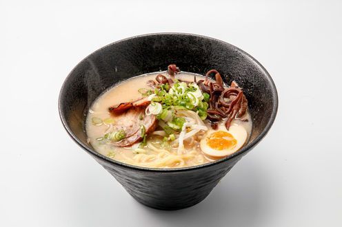 Black bowl of ramen with broth, noodles, sliced pork, egg, and green onions on a white background