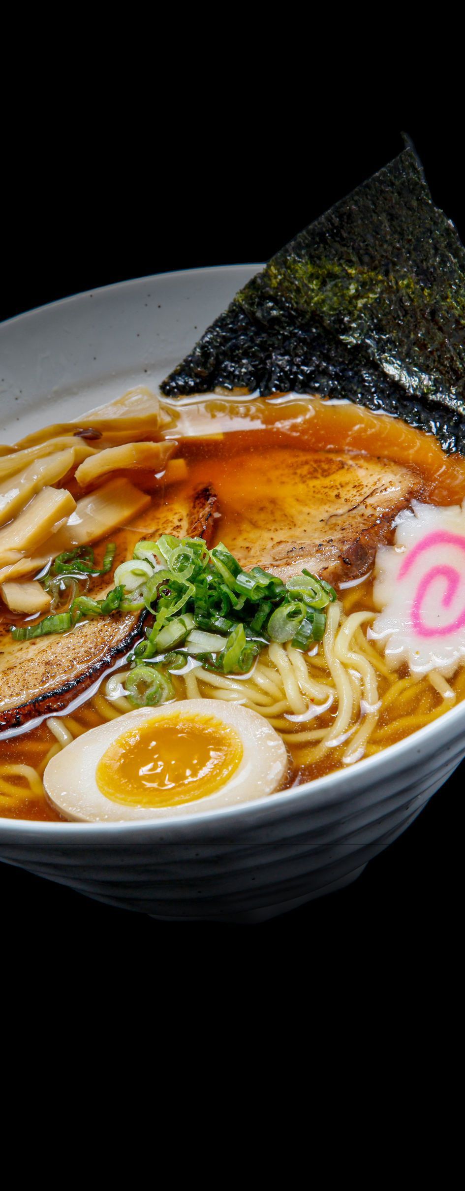 Bowl of ramen with broth, noodles, sliced pork, egg, green onion, and nori on a black background