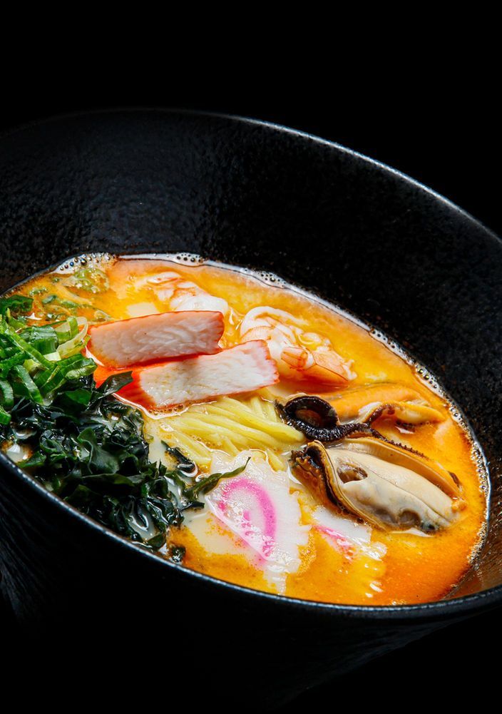 Japanese ramen in a black bowl with pork slices, egg, mushrooms, greens, and bright orange broth
