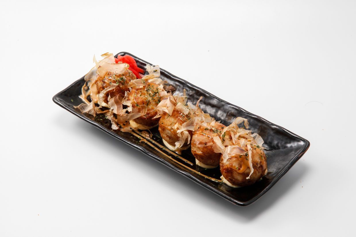 Black rectangular plate of takoyaki topped with bonito flakes and sauce on a white background