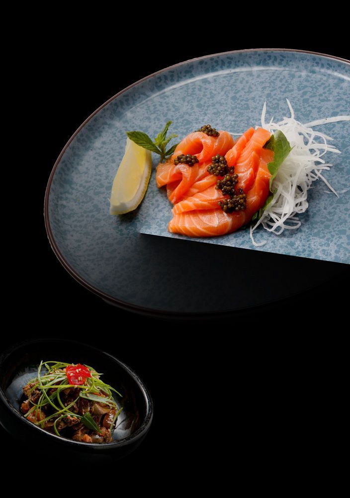 Salmon sashimi on a blue plate with garnish, beside a small bowl of greens and vegetables