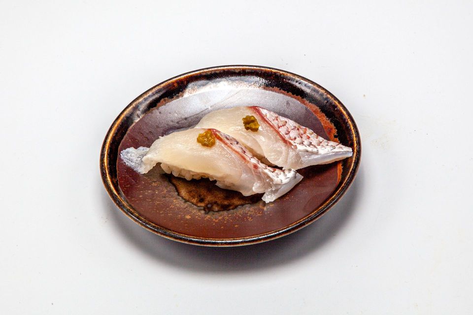 Two sushi pieces on a brown plate, topped with glossy sauce and garnishes