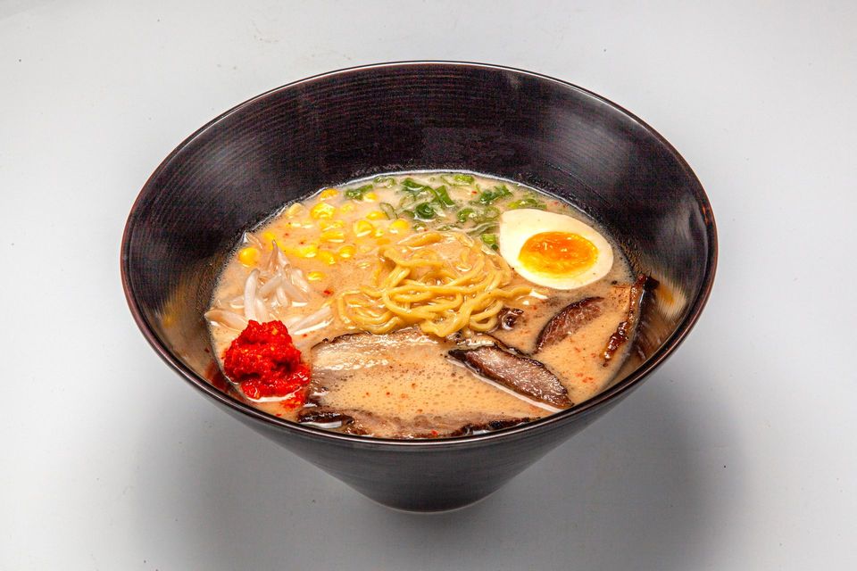 Black bowl of ramen with egg, noodles, mushrooms, and red garnish on a white background