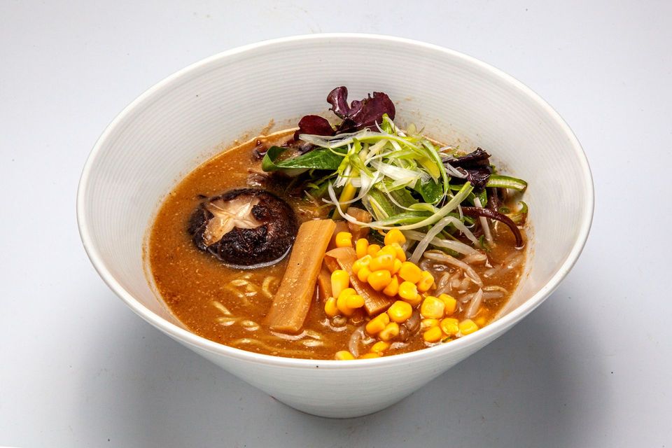 Bowl of ramen with corn, mushrooms, and greens in a light broth on a white background