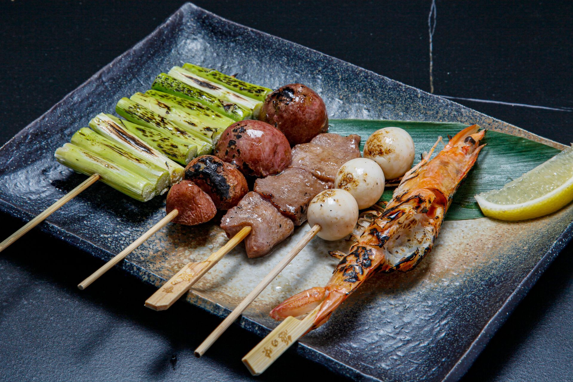Grilled skewers with assorted meats and vegetables on a black plate, garnished with lemon wedge