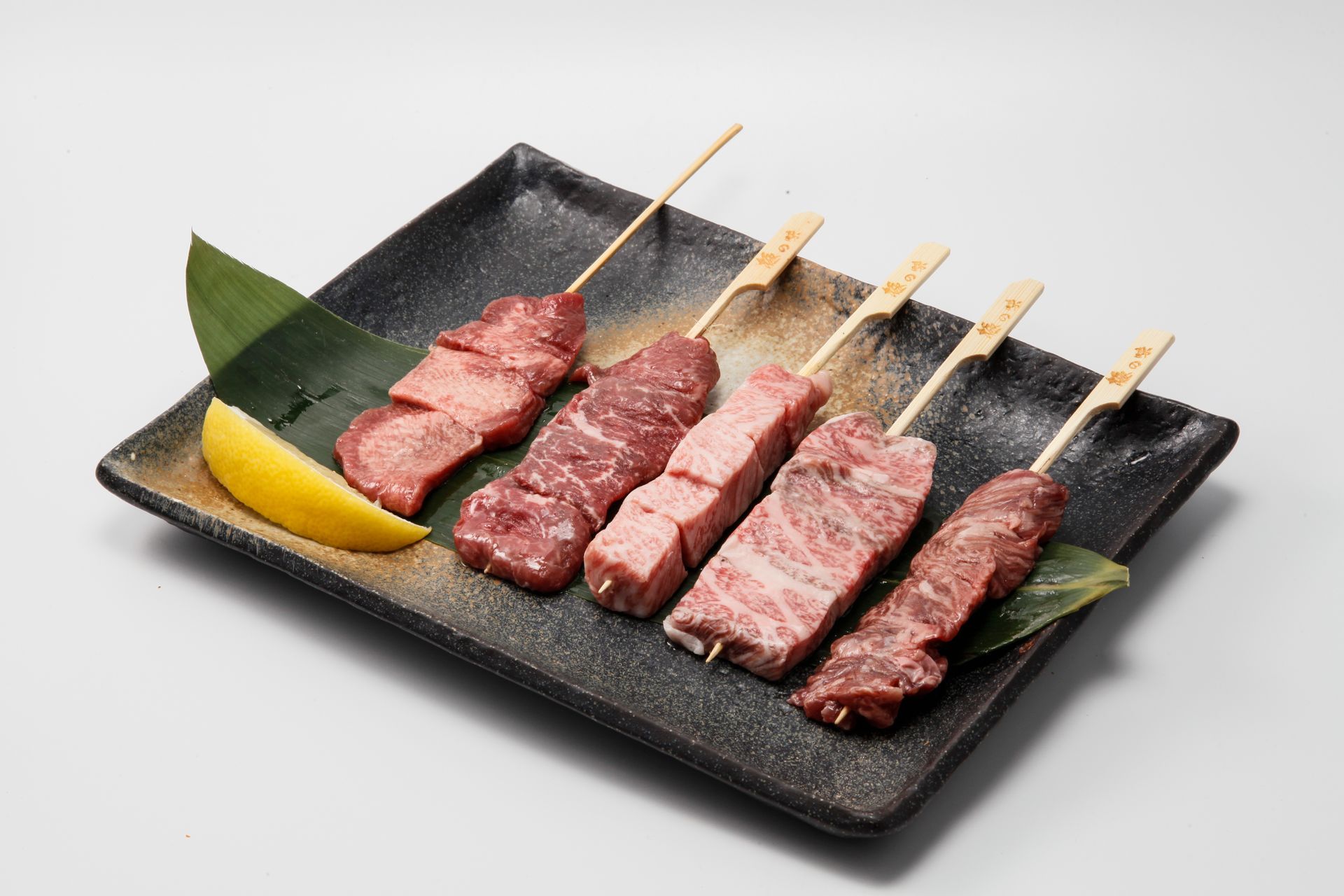 Grilled beef skewers on a black plate with a lemon wedge and green leaf garnish
