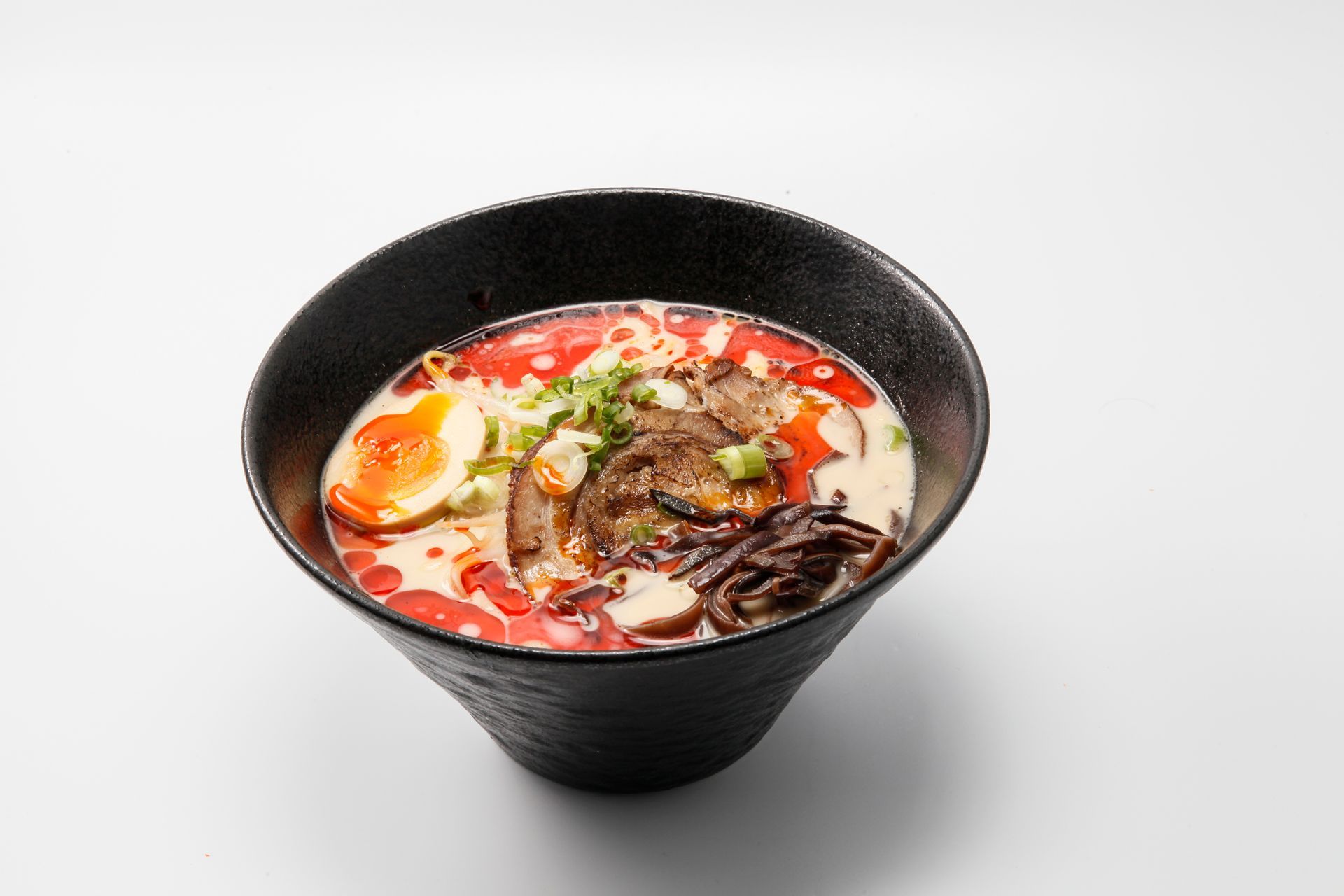 Black bowl of ramen topped with egg, meat, mushrooms, and red broth on a white background