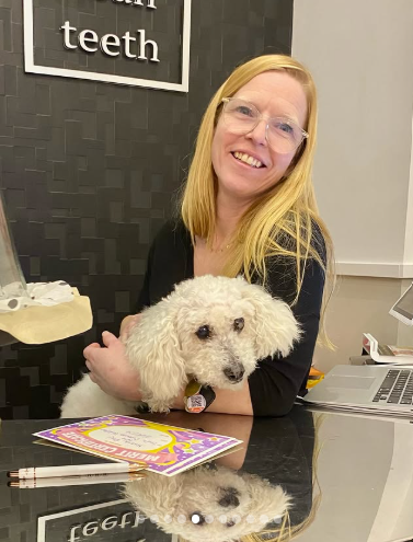 Woman smiling with a fluffy white dog at a desk in a dental office
