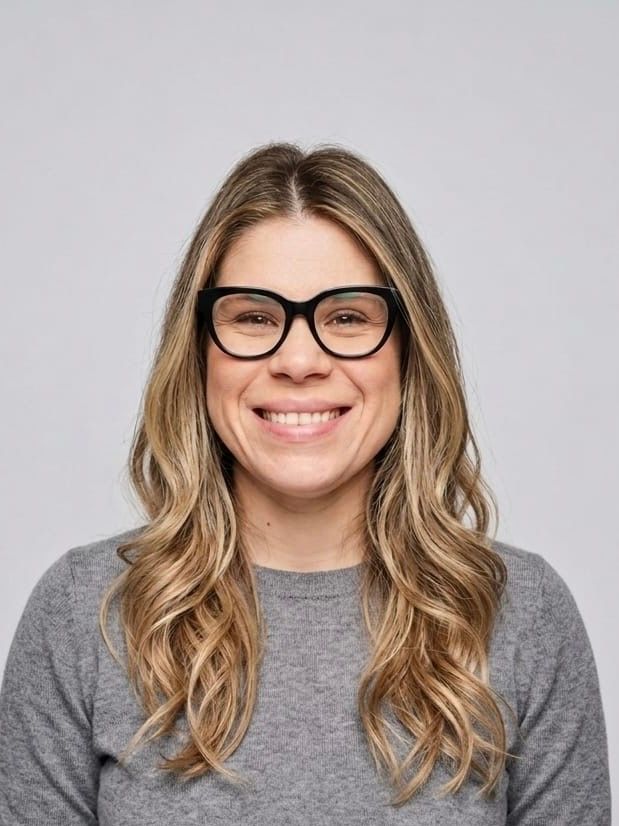 Smiling person with long wavy hair and black glasses wearing a gray sweater on a light background