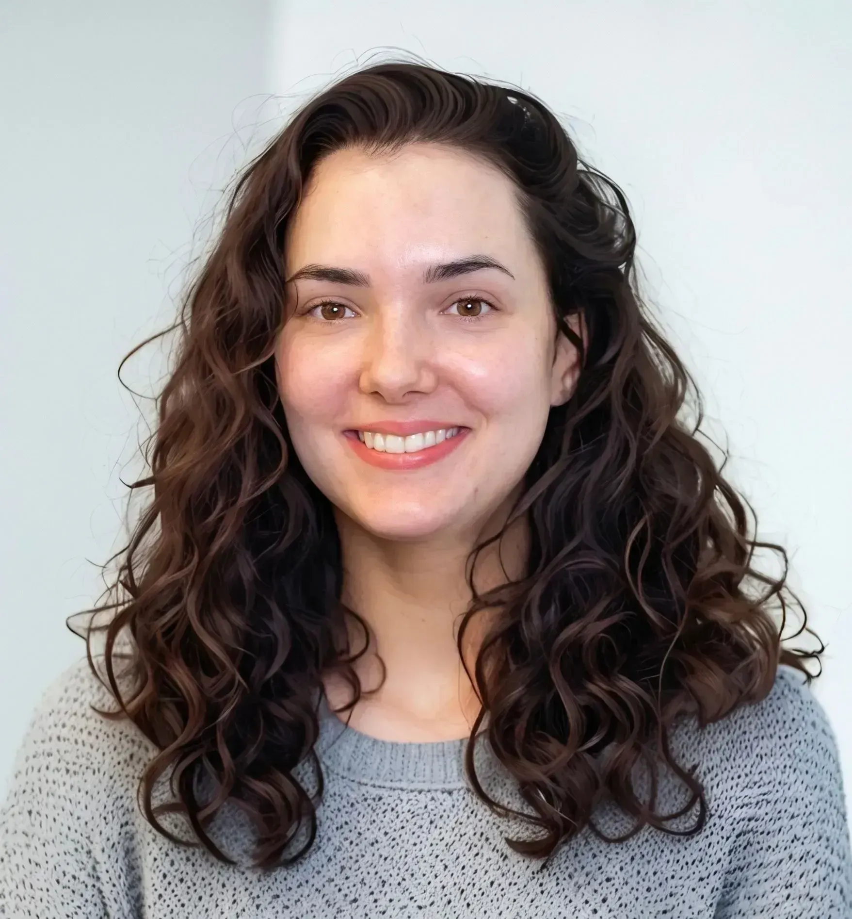 Smiling person with curly dark hair wearing a gray sweater against a light background