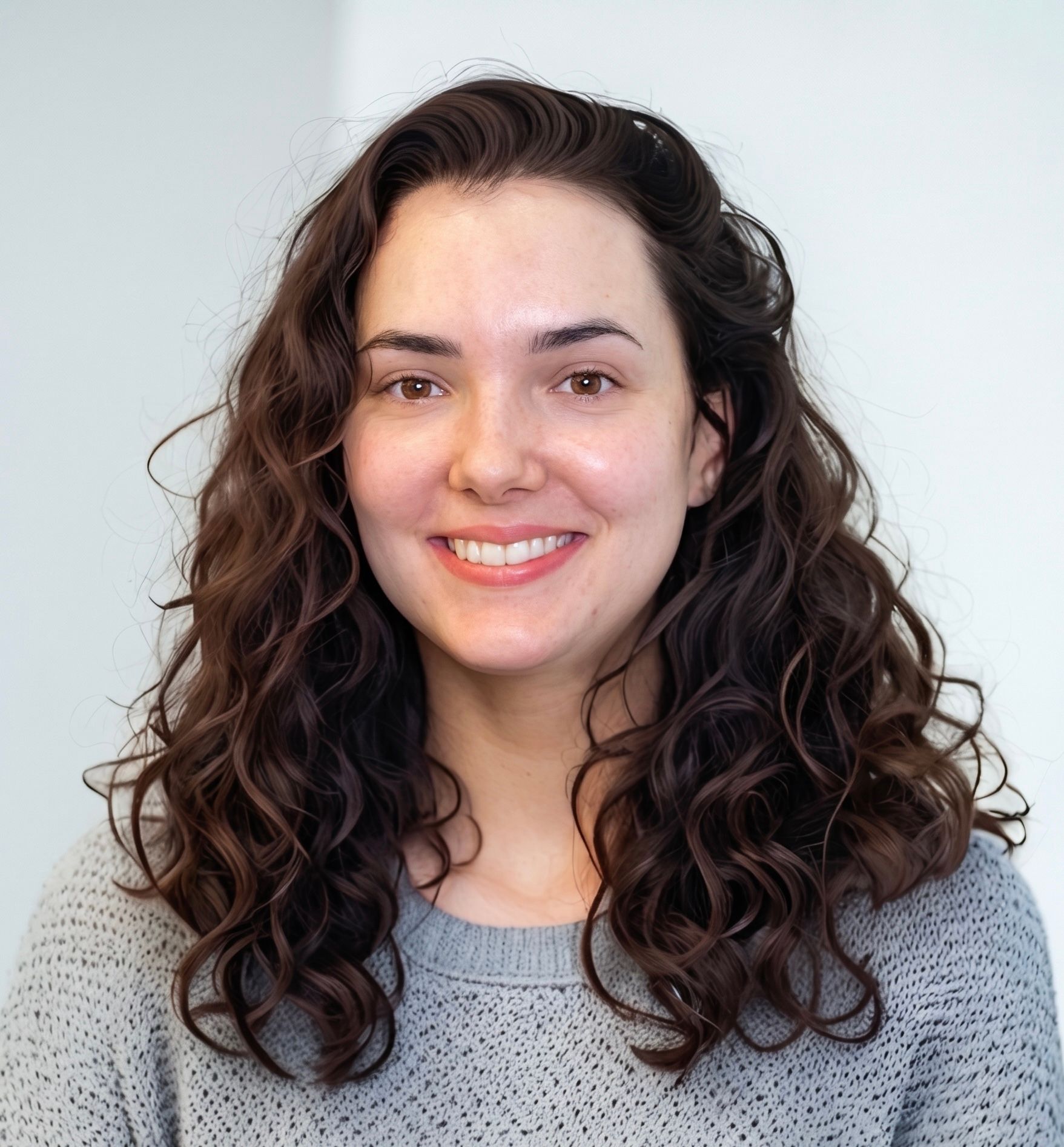 Smiling person with curly dark hair wearing a gray sweater against a light background