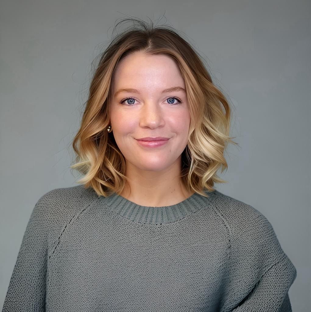 Smiling person with shoulder-length wavy blonde hair in a gray sweater against a plain gray background