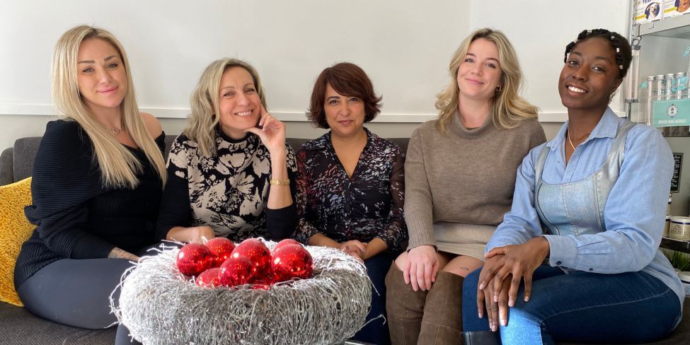 Five women sit on a couch in a bright living room, smiling beside a bowl of red flowers.