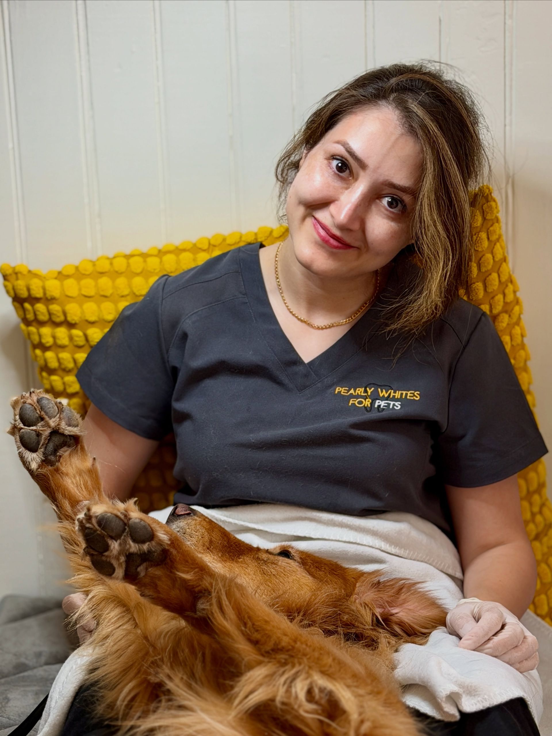 Woman in dark scrubs smiling on a yellow couch with a fluffy dog resting on her lap.