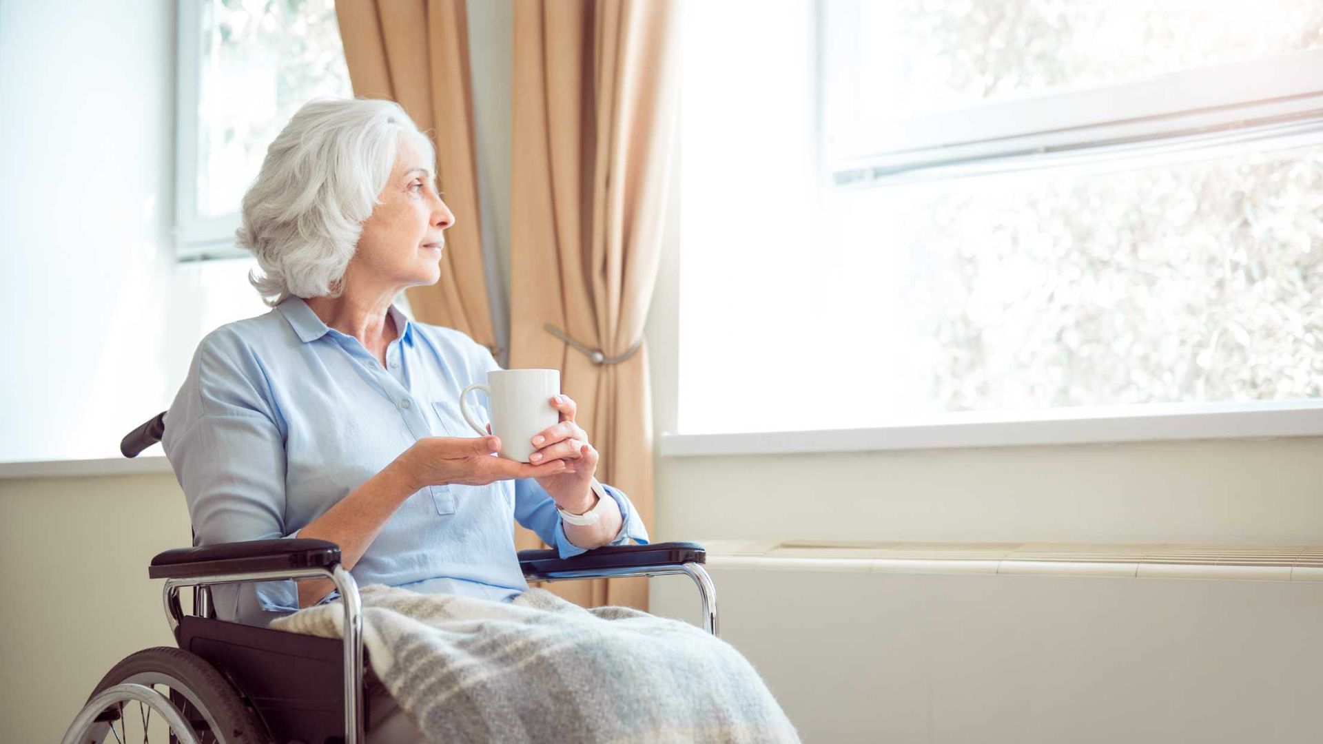 Elderly woman in wheelchair looking out window, holding a mug.