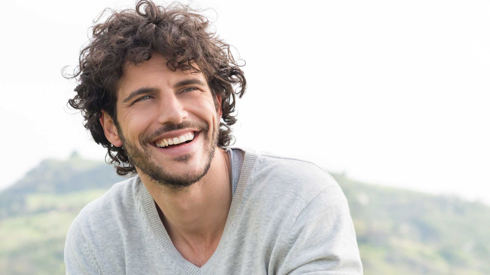 Man with curly hair smiles broadly outdoors, wearing a light gray sweater.