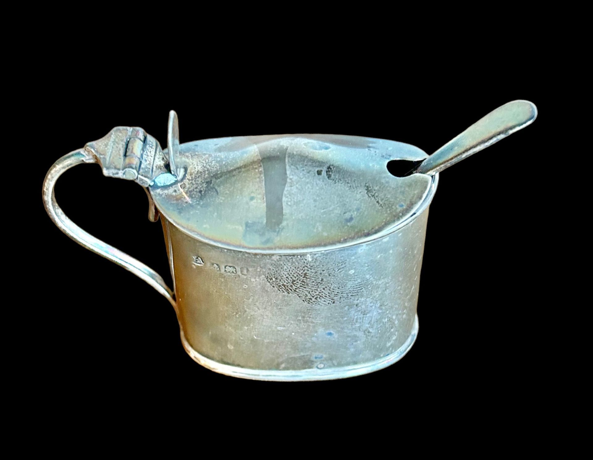 Een ovale, verweerde metalen mosterdpot met deksel en bijpassende lepel, geplaatst tegen een effen zwarte achtergrond.