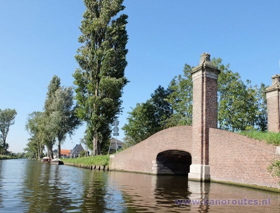 Op een zonnige dag overspant een bakstenen boogbrug een rustig kanaal, geflankeerd door hoge populieren en groen gras.