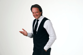 Een persoon, gekleed in een donker vest, wit overhemd en stropdas, glimlacht en gebaart met zijn hand tegen een witte achtergrond.